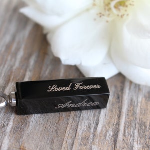 May include: Black rectangular cremation jewelry pendant with a silver loop. The pendant is engraved with the words "Loved Forever" and "Andrea".