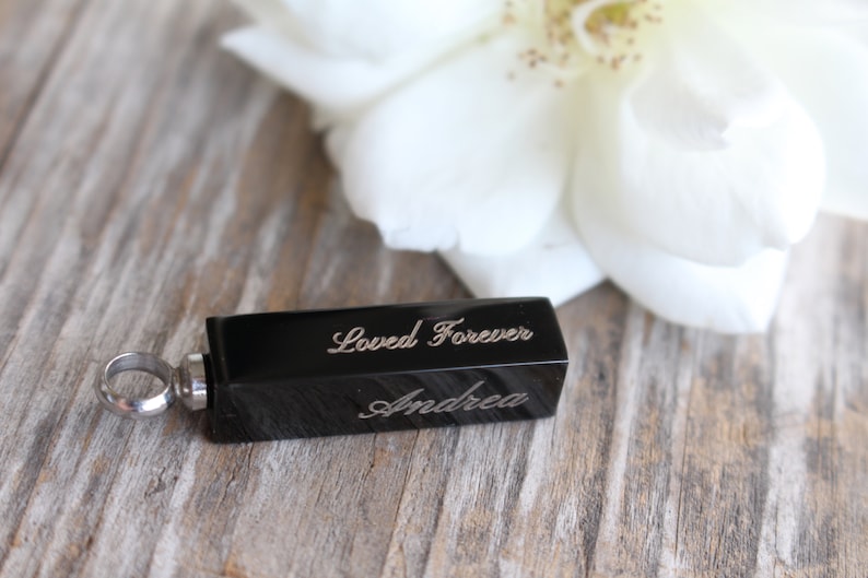 May include: Black rectangular cremation jewelry pendant with a silver loop. The pendant is engraved with the words "Loved Forever" and "Andrea".