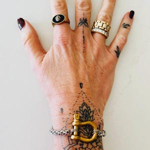 Pu&ograve; includere: Un braccialetto dorato con una grande chiusura a forma di D. Il braccialetto &egrave; al polso di una persona con un tatuaggio in bianco e nero di un disegno di mandala. La persona indossa un anello d'oro con una pietra nera al dito.