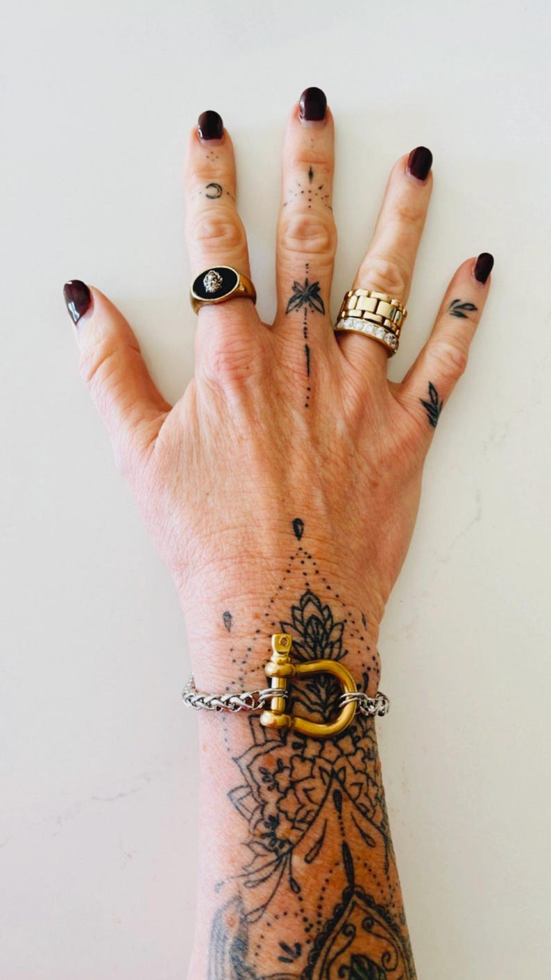Pu&ograve; includere: Un braccialetto dorato con una grande chiusura a forma di D. Il braccialetto &egrave; al polso di una persona con un tatuaggio in bianco e nero di un disegno di mandala. La persona indossa un anello d'oro con una pietra nera al dito.