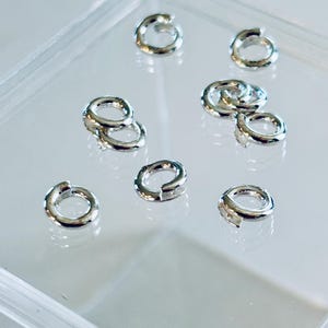 Pu&ograve; includere: Primo piano di un gruppo di anelli di salto in argento in un contenitore di plastica trasparente. Gli anelli di salto sono di diverse dimensioni e vengono utilizzati per la creazione di gioielli.