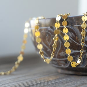 May include: A delicate gold-coloured necklace with small, round, flat charms. The necklace is draped over the edge of a carved, dark brown bowl. The background is a blurred, neutral colour.