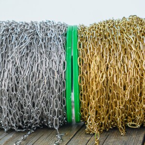 May include: Two spools of chain, one silver and one gold, are wound around green plastic spools. The spools are sitting on a wooden surface.