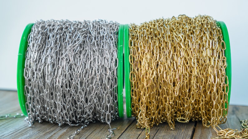 May include: Two spools of chain, one silver and one gold, are wound around green plastic spools. The spools are sitting on a wooden surface.