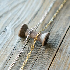 May include: Two chains of different metals, one silver and one gold, are draped over a wooden spool. The chains are both oval-linked and are lying on a wooden surface.