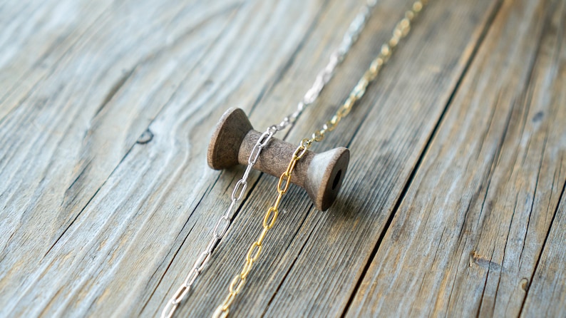 May include: Two chains of different metals, one silver and one gold, are draped over a wooden spool. The chains are both oval-linked and are lying on a wooden surface.