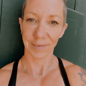 May include: A woman with short, blonde hair is wearing a black tank top and a gold chain necklace. She is standing in front of a green wall.