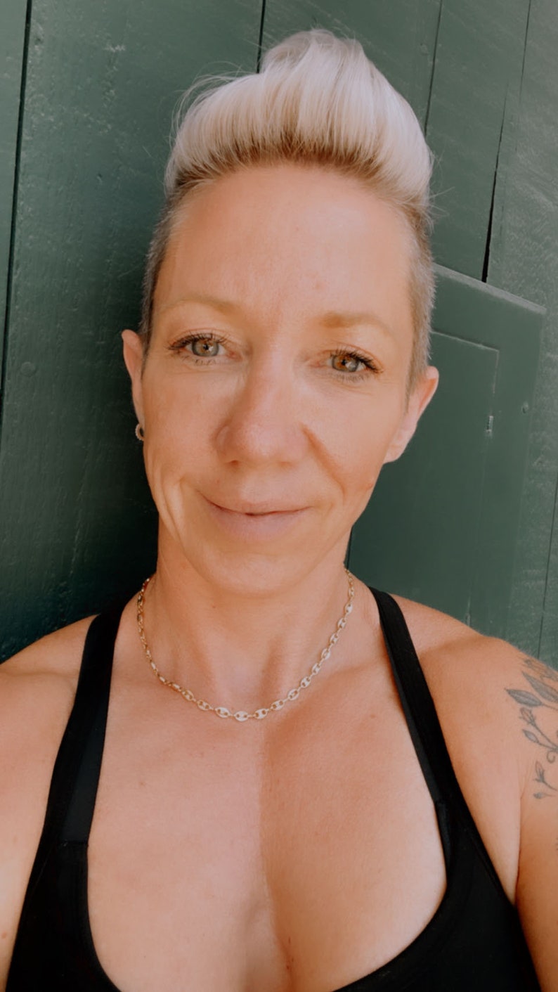 May include: A woman with short, blonde hair is wearing a black tank top and a gold chain necklace. She is standing in front of a green wall.