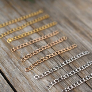 May include: A set of six chain links in different finishes, including gold, rose gold, and silver. The chains are laid out on a wooden surface.