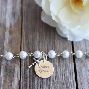 May include: A white pearl bracelet with a silver charm that says "Quinn Kendall" on a wooden background with a white rose.