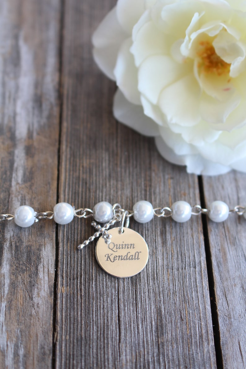May include: A white pearl bracelet with a silver charm that says "Quinn Kendall" on a wooden background with a white rose.
