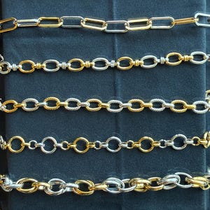 May include: Close-up of several gold and silver chain necklaces. The chains feature various link styles, including oval, rectangular, and textured designs. The jewelry is displayed against a dark background, highlighting the metallic shine.