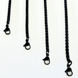 May include: Close-up of several black chain necklaces with lobster claw clasps against a white background. The chains are made of small, interconnected links. The clasps are attached to the ends of the chains.