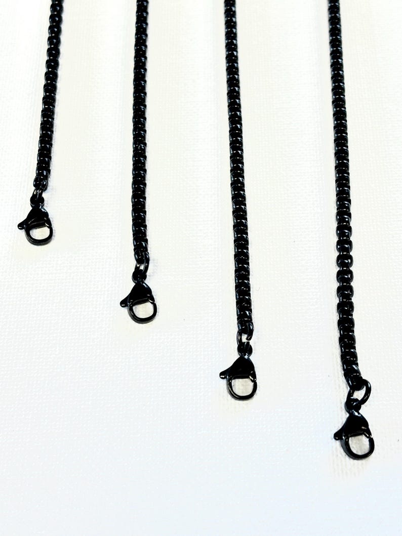 May include: Close-up of several black chain necklaces with lobster claw clasps against a white background. The chains are made of small, interconnected links. The clasps are attached to the ends of the chains.