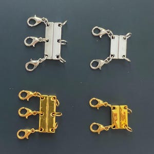 May include: Four multi-strand necklace clasps. Two are silver-toned, and two are gold-toned. Each clasp has three lobster claw clasps attached to a rectangular bar with a magnetic closure. The clasps are on a dark grey surface.