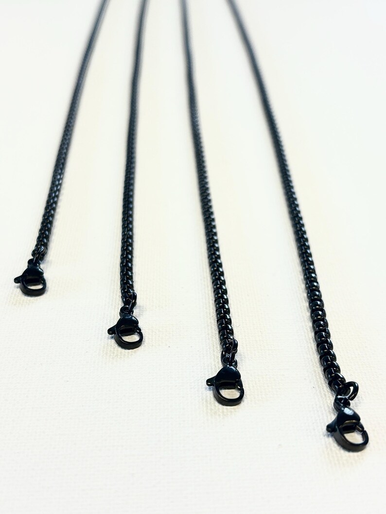 May include: Four black chain necklaces with lobster claw clasps are displayed against a white backdrop. The chains are evenly spaced and angled, creating a sense of depth. These chains are likely for jewellery making or as necklace components.