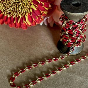May include: A spool of red and gold chain with a red enamel finish. The chain is on a wooden spool and is surrounded by red and pink flowers.