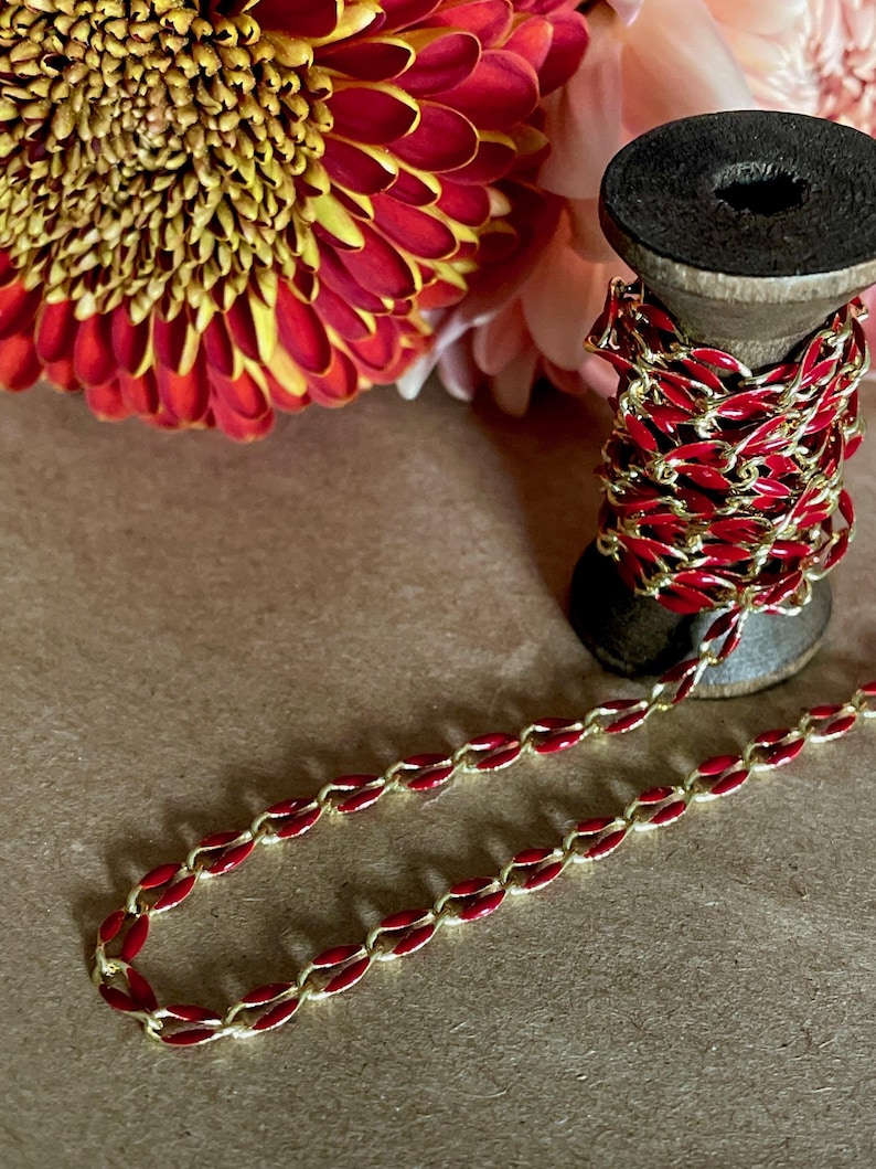 May include: A spool of red and gold chain with a red enamel finish. The chain is on a wooden spool and is surrounded by red and pink flowers.