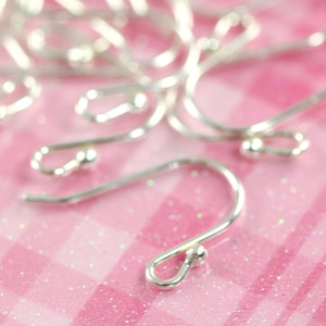 May include: A close-up of a pile of silver earring hooks on a pink glittery surface. The hooks are made of metal and have a small ball at the end.