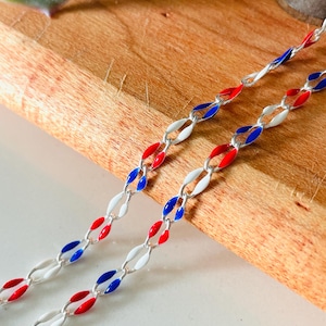 May include: Two strands of a silver chain with red, white, and blue enamel links. The links are oval-shaped and arranged in a repeating pattern. The chains are displayed on a wooden surface, with a blurred background.