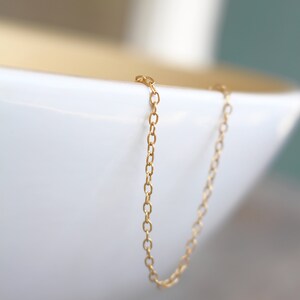 May include: A gold chain necklace with a delicate, simple design. The chain is hanging over the edge of a white bowl.
