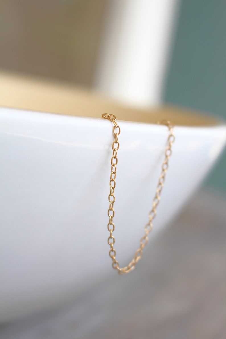 May include: A gold chain necklace with a delicate, simple design. The chain is hanging over the edge of a white bowl.