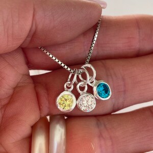 May include: A silver chain necklace with three charms, each featuring a different colored gemstone. The charms are a yellow, a clear, and a blue gemstone.