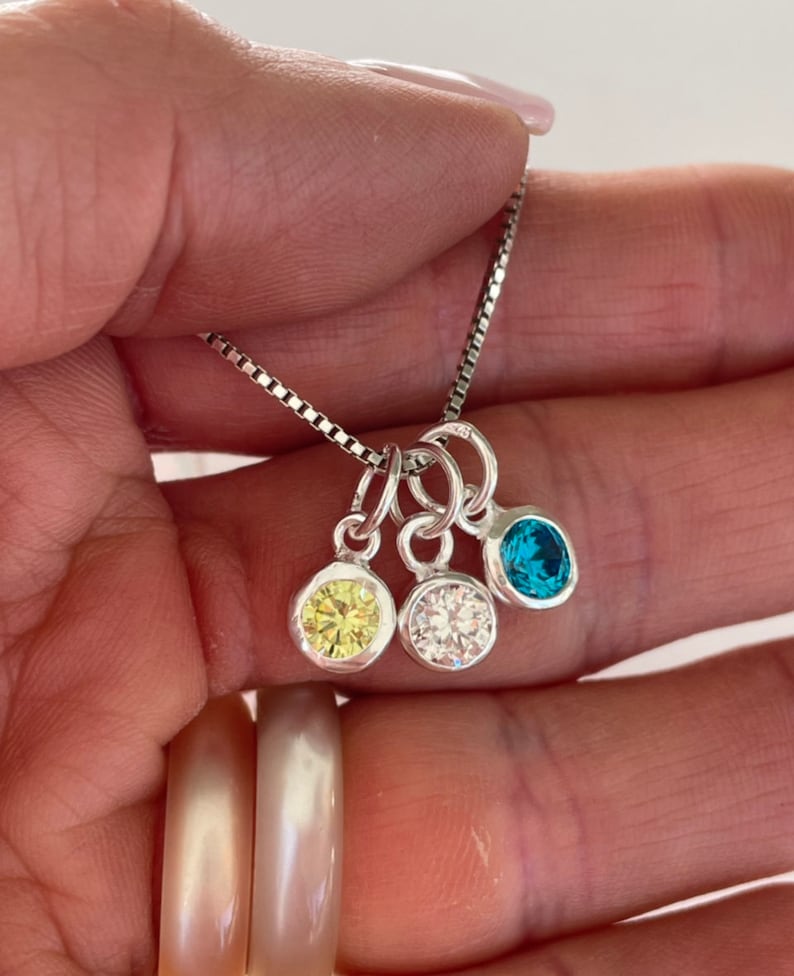 May include: A silver chain necklace with three charms, each featuring a different colored gemstone. The charms are a yellow, a clear, and a blue gemstone.