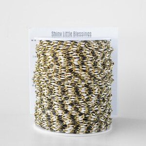 May include: A spool of gold and black chain trim with the words "Shiny Little Blessings" printed on the label.