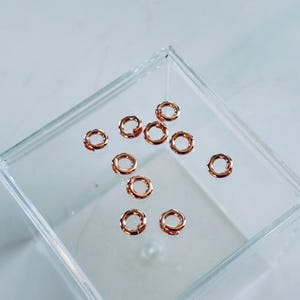 Pu&ograve; includere: Primo piano di un contenitore di plastica trasparente pieno di piccoli anelli di salto dorati. Gli anelli sono disposti in modo casuale.