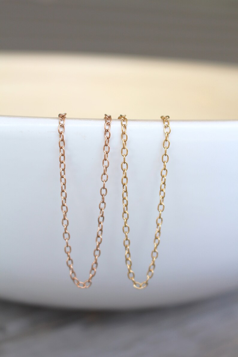 May include: Two delicate gold chains, one in rose gold and one in yellow gold, are displayed against a white background. The chains are both simple and elegant, making them perfect for layering or wearing alone.