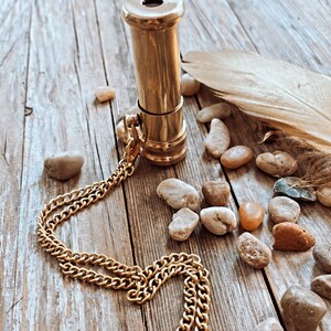 K&ouml;nnte beinhalten: Eine goldfarbene Teleskop-Anh&auml;nger-Halskette aus Messing mit einer Kette. Die Kette liegt auf einer Holzoberfl&auml;che mit kleinen Steinen, die um sie herum verstreut sind.