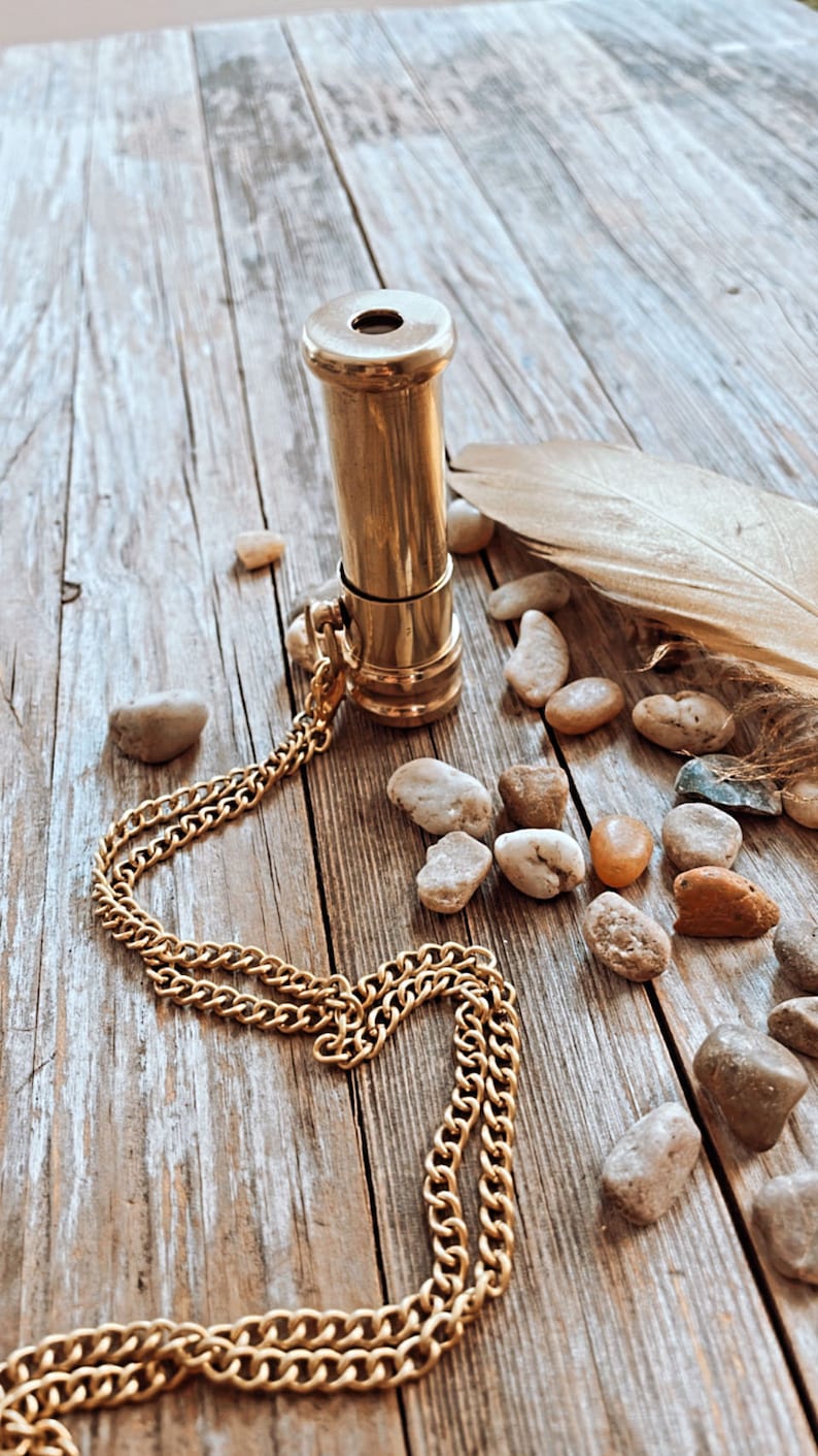 K&ouml;nnte beinhalten: Eine goldfarbene Teleskop-Anh&auml;nger-Halskette aus Messing mit einer Kette. Die Kette liegt auf einer Holzoberfl&auml;che mit kleinen Steinen, die um sie herum verstreut sind.