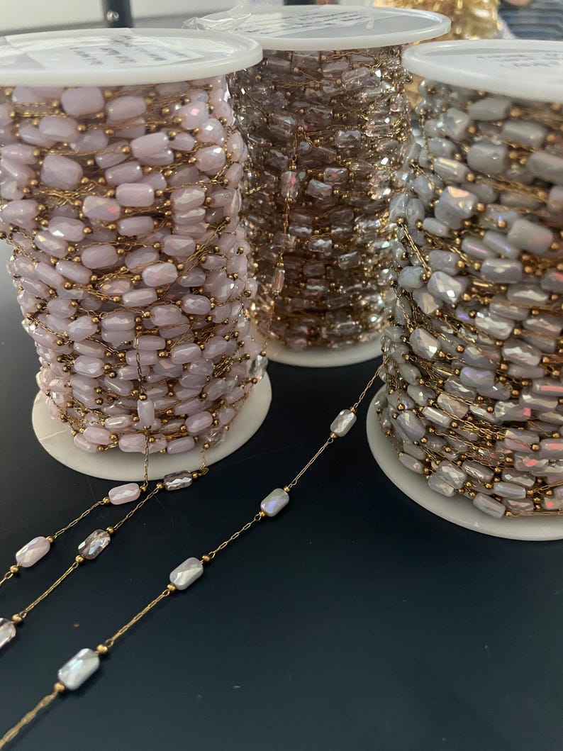 May include: Spools of beaded chain in three colors: pale pink, bronze, and iridescent white. The beads are rectangular and strung on gold-colored wire. The spools are on white cardboard spools.