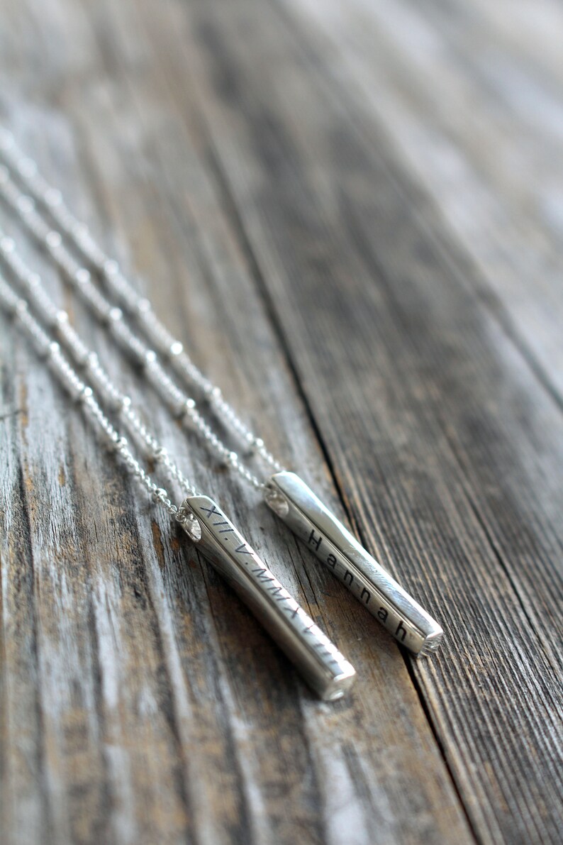 May include: Two silver necklaces with personalized bar pendants. One pendant reads "Hannah" and the other reads "XI.V.MMXVII".