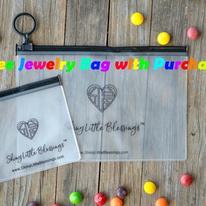 May include: Two clear plastic zippered bags with a black zipper and a black ring pull. The bags are labeled "Shiny Little Blessings" and feature a heart design. The text "Free Jewelry Bag with Purchase" is written in rainbow letters across the top of the image.