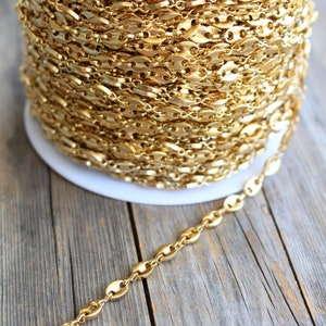 Op de afbeelding: Een spoel met een gouden ketting met een marinierslink-patroon. De ketting is op een witte spoel gewikkeld en een deel van de ketting ligt op een houten oppervlak.