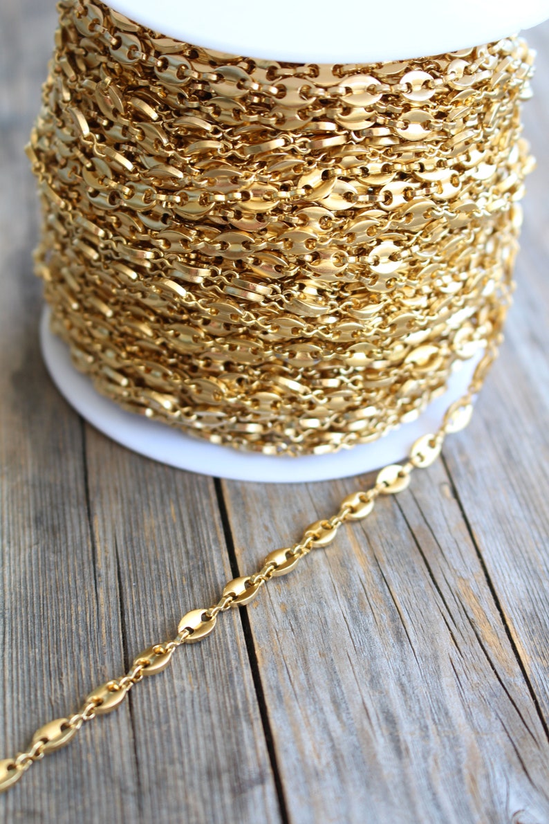 Op de afbeelding: Een spoel met een gouden ketting met een marinierslink-patroon. De ketting is op een witte spoel gewikkeld en een deel van de ketting ligt op een houten oppervlak.