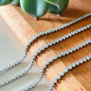 May include: Close-up of three silver tennis bracelets with clear, sparkling stones. The bracelets are arranged diagonally across a wooden surface, with a succulent plant in the background. The jewelry is displayed in a bright, well-lit setting.