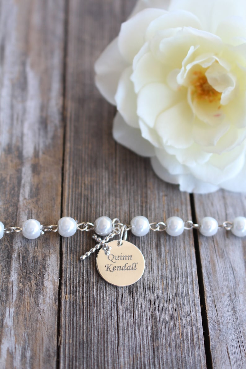 May include: A white pearl bracelet with a silver charm that says "Quinn Kendall" on a wooden background with a white rose.