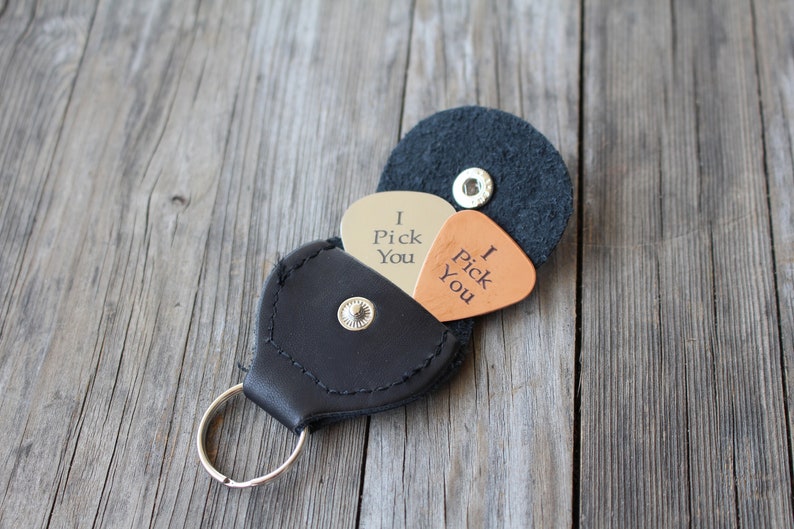 May include: A black leather keychain with a guitar pick holder. The holder has two guitar picks inside, one silver and one brown. The silver pick says "I Pick You" and the brown pick says "I Pick You".