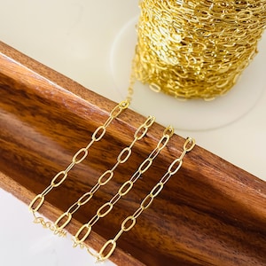 May include: Gold-colored chain in a paperclip style, displayed on a wooden tray. A spool of the same chain is visible in the background. The chain links are oval-shaped and evenly spaced, creating a delicate and elegant appearance. Ideal for jewelry making.