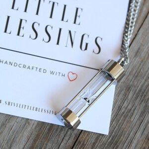 May include: A silver necklace with a small hourglass pendant. The pendant is clear glass with a silver top and bottom. The necklace is on a white card with the text "Shiny Little Blessings" and "Handcrafted with &hearts;".