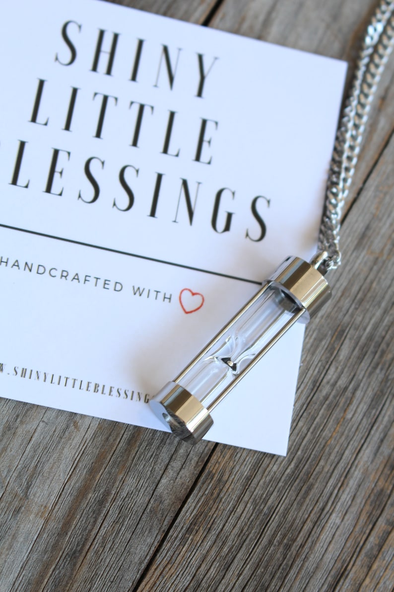 May include: A silver necklace with a small hourglass pendant. The pendant is clear glass with a silver top and bottom. The necklace is on a white card with the text "Shiny Little Blessings" and "Handcrafted with &hearts;".