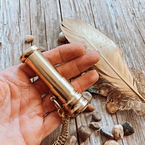 K&ouml;nnte beinhalten: Ein goldfarbener Metallr&ouml;hrenanh&auml;nger mit einer Kette. Der Anh&auml;nger liegt auf einer Holzoberfl&auml;che mit kleinen Steinen und einer Feder.