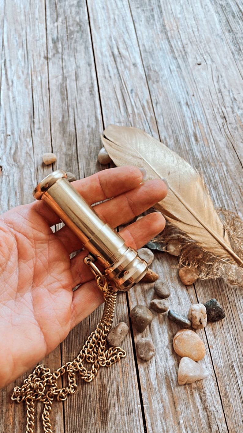 K&ouml;nnte beinhalten: Ein goldfarbener Metallr&ouml;hrenanh&auml;nger mit einer Kette. Der Anh&auml;nger liegt auf einer Holzoberfl&auml;che mit kleinen Steinen und einer Feder.
