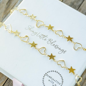 May include: A gold chain necklace with alternating star and heart charms. The necklace is displayed on a white box with the text "Shiny Little Blessings" and "Handcrafted with Love".