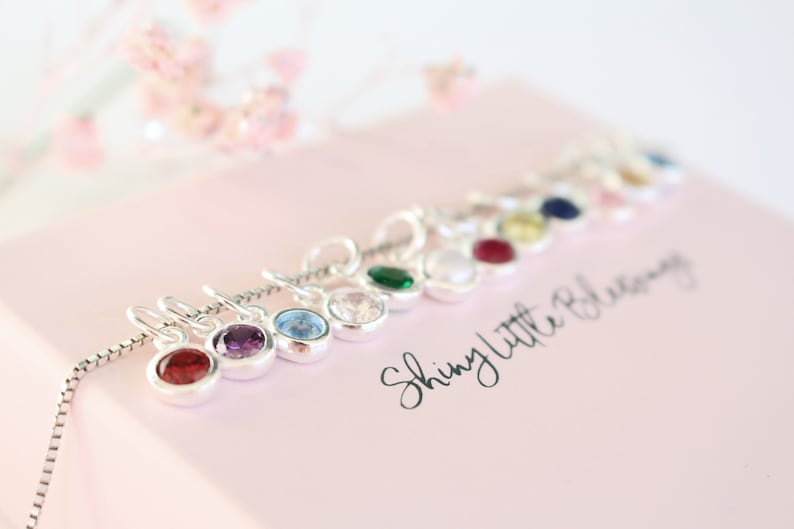 May include: A silver chain necklace with small charms featuring different colored gemstones. The charms are arranged in a row and the text "Shiny Little Blessings" is written on a pink surface.