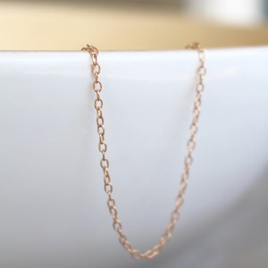 May include: A delicate rose gold chain necklace, hanging in a white bowl. The chain is made of small, oval-shaped links.