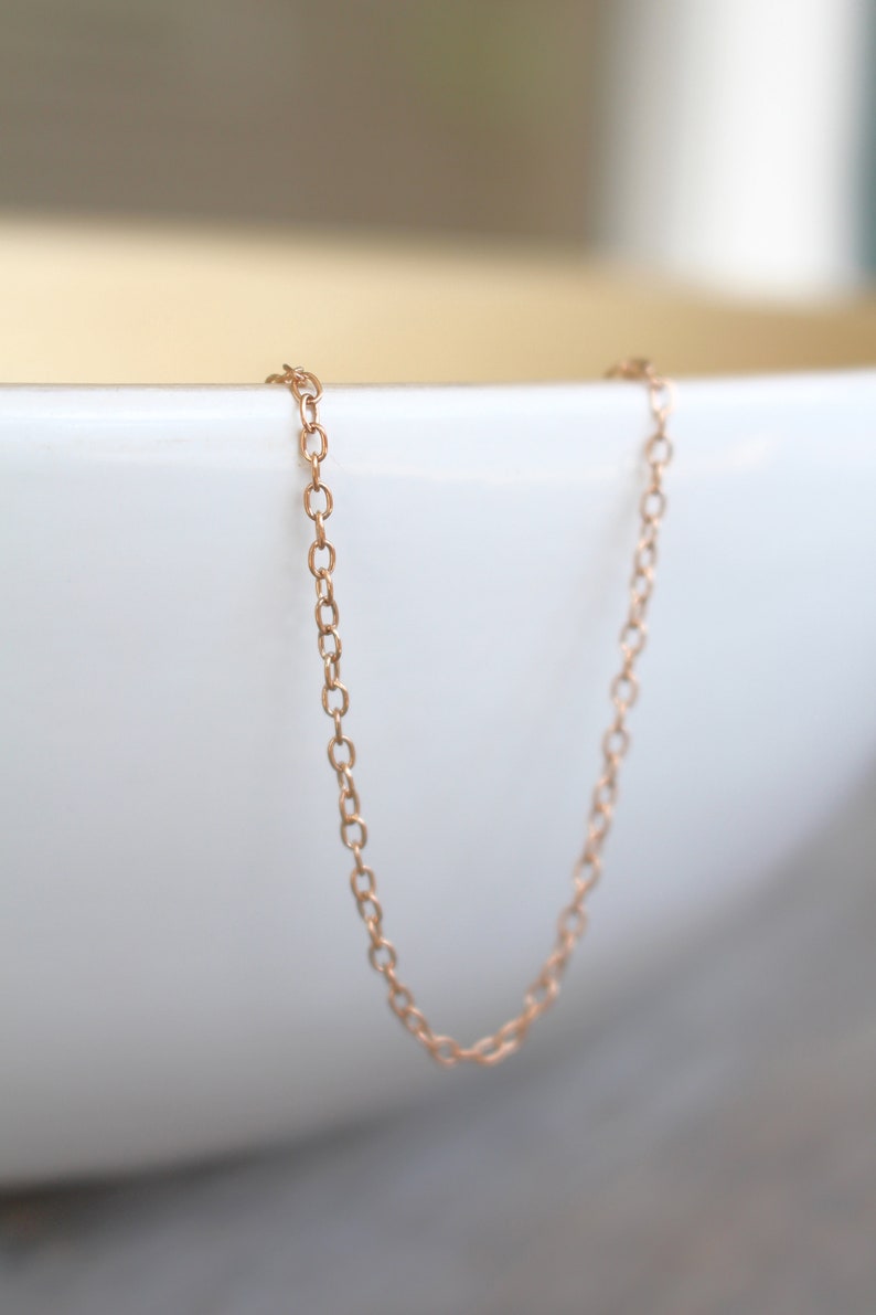 May include: A delicate rose gold chain necklace, hanging in a white bowl. The chain is made of small, oval-shaped links.